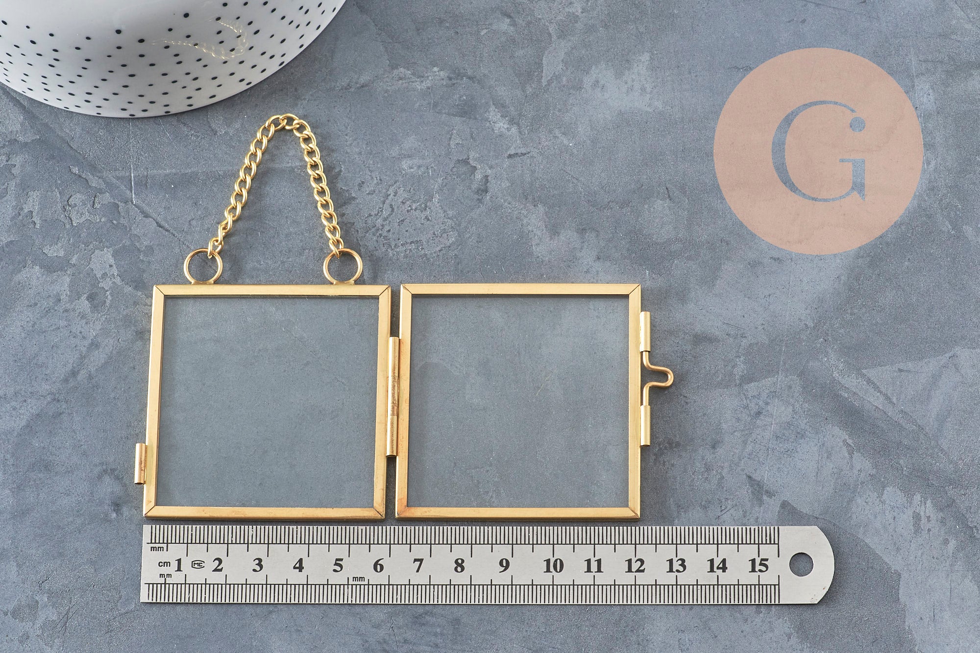 Cadre photo double articulé en verre et métal doré avec chaînette de suspension, posé sur un fond gris texturé.